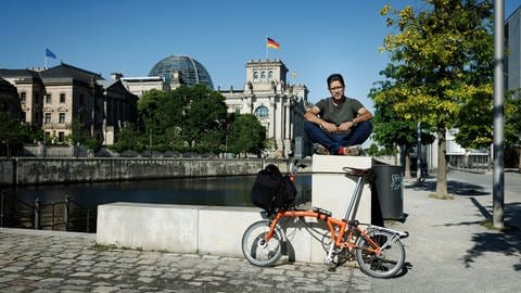 Journalist und Autor, Hasnain Kazim sitzt im Schneidersitz auf einer Mauer., daran lehnt sein orangefarbenes Fahrrad.