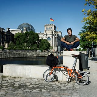 Journalist und Autor, Hasnain Kazim sitzt im Schneidersitz auf einer Mauer., daran lehnt sein orangefarbenes Fahrrad.