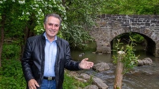 Naturschützer Claus-Peter Hutter zeigt mit der Hand auf einem Bach mit einer Steinbrücke.