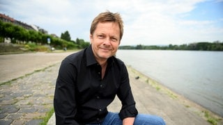 Martin Hecht, Politikwissenschaftler, sitzt mit aufgestützten Armen auf einer Mauer an einem Fluss.