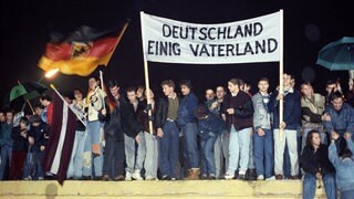 Jahrzehnte nach der Wiedervereinigung ist Deutschland gespalten, sagt Germanist Dirk Oschmann: Westdeutschland blicke auf Ostdeutschland, die ehemalige DDR, herab. Dabei fing alles so gut an: Mit der Deutschlandfahne und einem Transparent "Deutschland Einig Vaterland" stehen zahlreiche Berliner am 22.12.1989 auf der Berliner Mauer am Brandenburger Tor. Das Tor ist seit diesem Tag - 28 Jahre nach dem Mauerbau - wieder zugänglich.