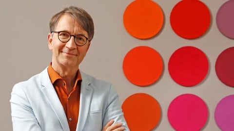 Prof. Axel Buether steht mit verschränkten Armen neben einer Wand mit roten und orangen Punkten. Prof. Axel Buether ist "Farbforscher": Seine Fachgebiete sind Farbpsychologie und Architektur. In SWR1 Leute erklärt er, wie uns Farben direkt oder indirekt bewegen und steuern.  