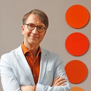 Prof. Axel Buether steht mit verschränkten Armen neben einer Wand mit roten und orangen Punkten. Prof. Axel Buether ist "Farbforscher": Seine Fachgebiete sind Farbpsychologie und Architektur. In SWR1 Leute erklärt er, wie uns Farben direkt oder indirekt bewegen und steuern.  