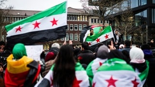 Feiernde schwenken die Flagge der syrischen Revolution vor dem Hauptbahnhof in Duisburg. Die Flagge der Gegner des Assad-Regimes zeigt drei horizontale Streifen - einen grünen, einen weißen und einen schwarzen - sowie drei rote Sterne.