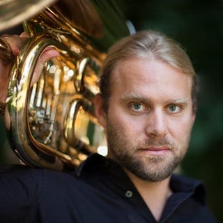 Andreas Martin Hofmeir, Tubist, Dirigent und Kabarettist, trägt seine Tuba auf der Schulter und blickt direkt in die Kamera.