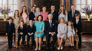 Das Familienfoto der schwedischen Königsfamile zum 50. Thronjibiläum von König Carl Gustaf von Schweden. Auf dem Foto zu sehen sind neben den drei Kindern des Königs und ihren Partnern auch seine Enkel und natürlich seine Frau Silvia. Sie tragen alle schicke Kleider oder Anzüge und stehen entsprechend angeordnet in einem königlichen Zimmer mit schönen Zierpflanzen.
