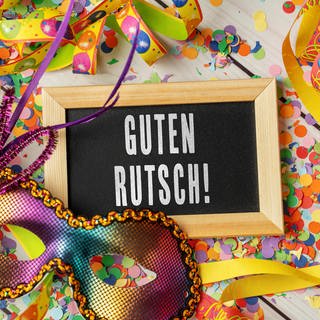 Silvesterparty Ideen: Auf einem Tisch sieht man Konfetti und Luftschlangen und eine glitzernde Maske. Dazischen liegt eine kleine Tafel, auf der geschrieben steht: Guten Rutsch.