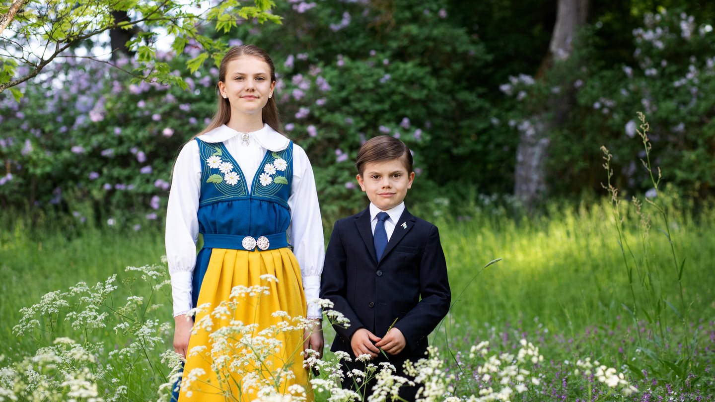 Prinzessin Estelle und Prinz Oscar posieren in traditioneller Tracht und Anzug auf einer grünen Wiese für den schwedischen Nationalfeiertag.
