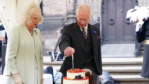 links steht Königin Camilla und schaut mit offenem Mund auf König Charles, der in Uniform rechts von ihr steht und eine Torte mit einem Messer vor sich auf dem Tisch anschneidet