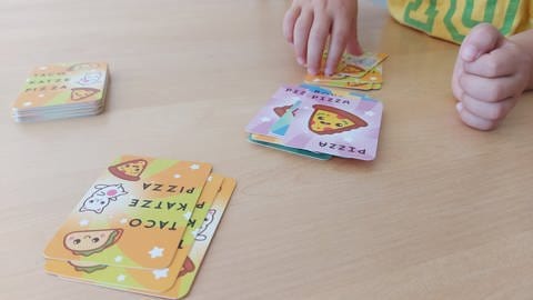Eine Kinderhand greift nach einer verdeckte Karte, um diese auf Stapel mit offen ausliegenden Karten zu legen: Die Jury "Spiel des Jahres" hat "Taco Katze Pizza Junior" zum "Kinderspiel des Jahres 2024" nominiert.