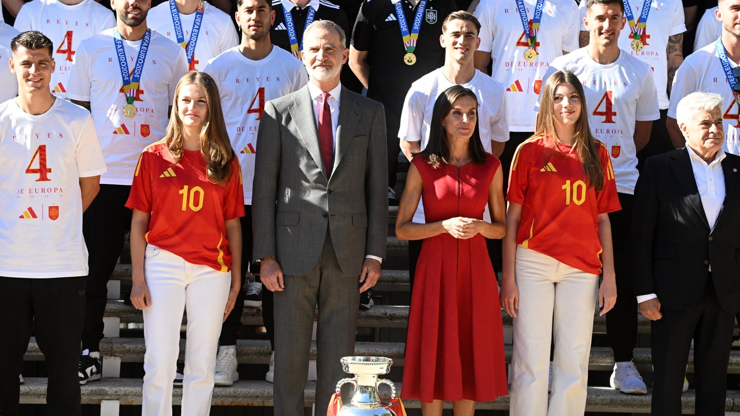 Die spanische Königsfamilie empfängt und posiert mit der spanischen Fußball-Nationalmannschaft nach deren EM-Sieg. Die Prinzessinnen Leonor und Sofia, sowie König Felipe und Königin Letizia stehen dabei in der Mitte direkt hinter dem Pokal. Um sie herum steht die Fußballmannschaft.