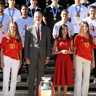Die spanische Königsfamilie empfängt und posiert mit der spanischen Fußball-Nationalmannschaft nach deren EM-Sieg. Die Prinzessinnen Leonor und Sofia, sowie König Felipe und Königin Letizia stehen dabei in der Mitte direkt hinter dem Pokal. Um sie herum steht die Fußballmannschaft.