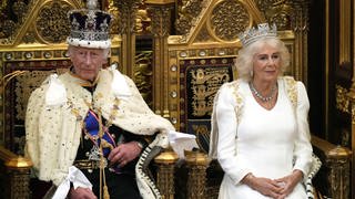 König Charles und seine Frau Königin Camilla sitzen auf einem prachtvollen Thron mit Kronen und Umhang und schicken Gewändern. Er wartet darauf seine Rede zur Parlamentseröffnung im House of Lords zu lesen, sie nahm soeben Platz neben ihm.
