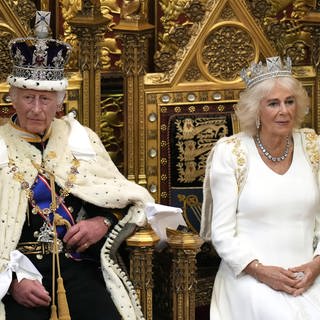 König Charles und seine Frau Königin Camilla sitzen auf einem prachtvollen Thron mit Kronen und Umhang und schicken Gewändern. Er wartet darauf seine Rede zur Parlamentseröffnung im House of Lords zu lesen, sie nahm soeben Platz neben ihm.