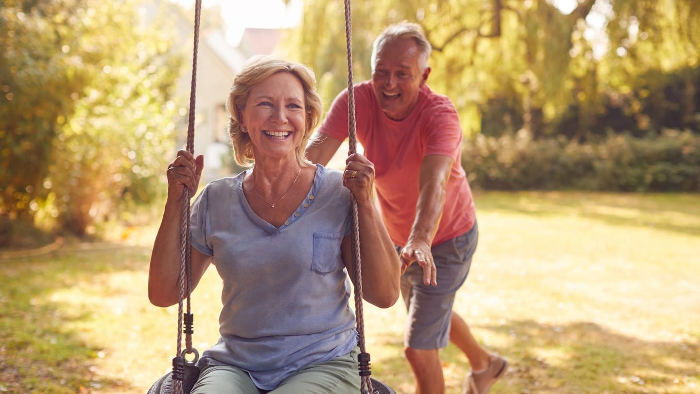 Ein Seniorin sitzt auf einer Schaukel, ein Senior schuckt sie an, beide lachen: Schaukeln macht auch Erwachsene glücklich. Es hilft dabei, gesund zu bleiben und erhöht das Wohlbefinden.