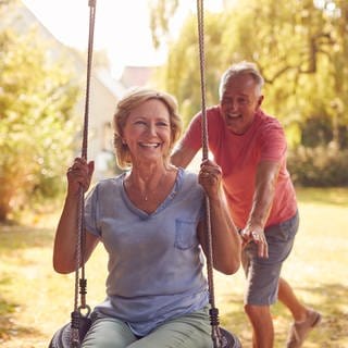 Ein Seniorin sitzt auf einer Schaukel, ein Senior schuckt sie an, beide lachen: Schaukeln macht auch Erwachsene glücklich. Es hilft dabei, gesund zu bleiben und erhöht das Wohlbefinden.