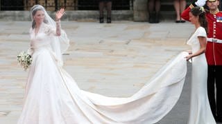 Royale Brautkleider – die Trends im britischen Königshaus: Prinzessin Kate trägt bei ihrer Hochzeit ein weißes Kleid und winkt. Die Schleppe hält ihre Schwester Pippa.