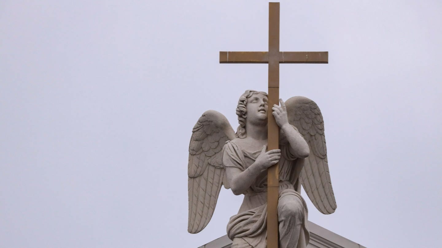 Kirchensymbole: Eine Engelsstatue aus hellgrauem Stein umklammert ein Holzkreuz auf dem Dach.