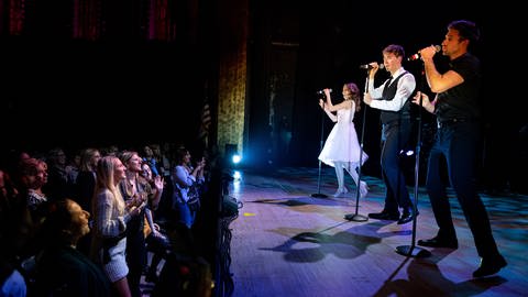 Drei Musicaldarsteller stehen vorne an der Bühne und singen, das Publikum klatscht und lacht.