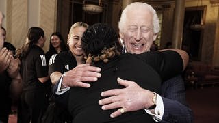 König Charles III. trifft die neuseeländische Rugby-Union-Mannschaft der Black Ferns im Buckingham Palace in London, umarmt eine Spielerin und hat dabei die Augen geschlossen.