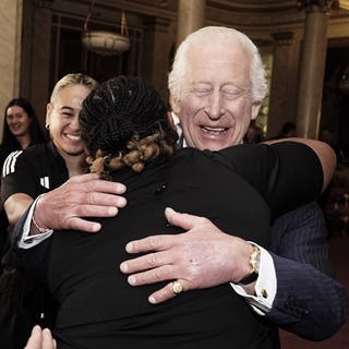 König Charles III. trifft die neuseeländische Rugby-Union-Mannschaft der Black Ferns im Buckingham Palace in London, umarmt eine Spielerin und hat dabei die Augen geschlossen.