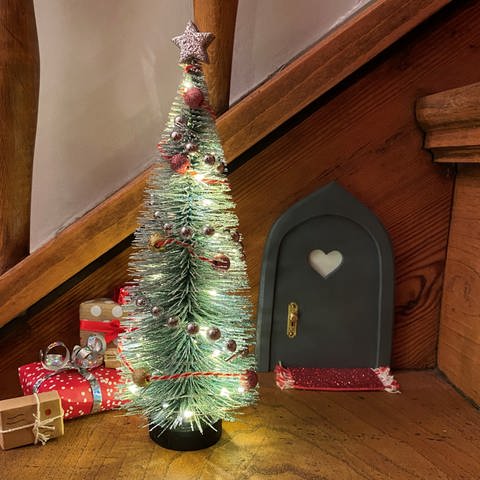 Trend für Weihnachten 2024: An einer Holztreppe ist im Advent eine kleine Wichteltür befestigt. Davor steht ein kleiner, geschmückter Deko-Weihnachtsbaum.