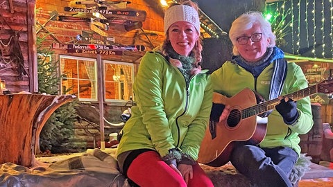 Annette Kienzle-Ehrlich und Werner Acker mit Gitarre von "Die kleinste SWR4 Band der Welt" sitzen auf einer Bank auf einem Weihnachtsmarkt.