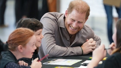 Prinz Harry sitzt an einem Tisch mit Kindern. Er trägt ein hellbraunes Hemd und beugt sich zu den Kindern nach unten und lächelt.