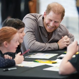 Prinz Harry sitzt an einem Tisch mit Kindern. Er trägt ein hellbraunes Hemd und beugt sich zu den Kindern nach unten und lächelt.