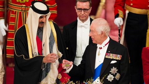 Links steht ein Scheich mit einem weißen Kopftuch und hält ein glas mit einer durchsichtigen Flüssigkeit in der Hand, er prostet dem Mann rechts, König Charles, im schwarzen Wrack mit Orden behängt zu, der auch ein Glas mit einer durchsichtigen Flüssigkeit in der Hand hält zu