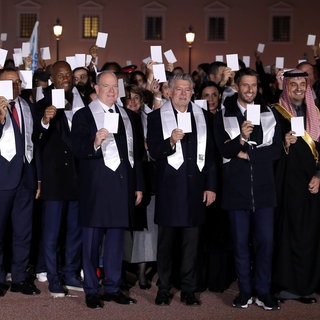 Ein Gruppenbild mit Albert, dem Fürsten von Monaco, der im schwarzen Anzug in der Mitte steht, alle Leute auf dem Bild halten einen weißen Zettel hoch und tragen eine weiße Stola um den Hals