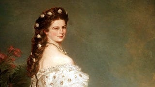 Sissi: Kaiserin Elisabeth hat helle Perlen in ihrem rot-braunen Haar, das zu einer aufwendigen Frisur gesteckt ist und trägt ein wunderschönes weißes Kleid mit goldenem Muster dazu.