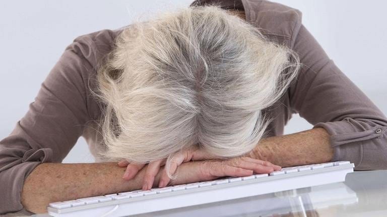 Eine grauhaarige Frau hat ihren Kopf müde auf die Tastatur ihres Computers gelegt.