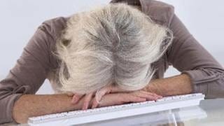 Eine grauhaarige Frau hat ihren Kopf müde auf die Tastatur ihres Computers gelegt.