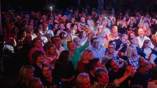 Eine bunt angeleuchtete Publikumsmenge hebt die Arme, lacht und schaut auf eine Bühne außerhalb des Bildes.