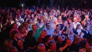 Eine bunt angeleuchtete Publikumsmenge hebt die Arme, lacht und schaut auf eine Bühne außerhalb des Bildes.