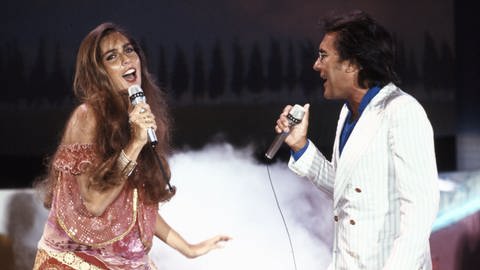 Romina Power singt mit langem offenen Haar und einem Glitzerkleid voll Freude in ein Mikrofon. Ihr gegenüber steht Al Bano, er singt ebenfalls mit ihr im Duo.