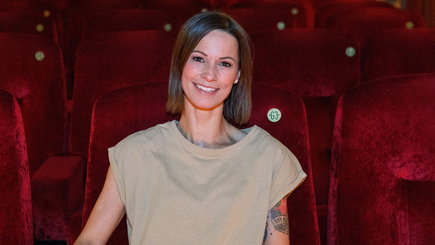 Sängerin Christina Stürmer trägt ein T-Shirt in der Farbe beige. Sie lächelt und sitzt in einem Kinosaal voller samtiger roter Sitze. An ihrem linken Arm sind Tattoos zu sehen.