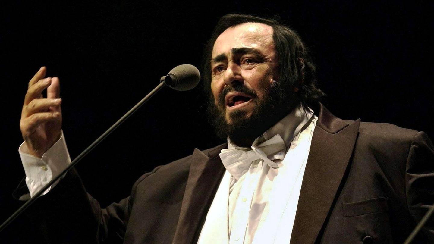 Der Opernsänger Luciano Pavarotti singt vor einem Mikrofon in einem schwarzen Frack.