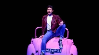 Das Hauptdarsteller des Musicals Grease sitzt auf einem rosa Auto und singt