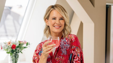 Monica Meier Ivancan steht mit einem Glas an der Hand in einem bunten Wickelkleid in der Küche an der Theke