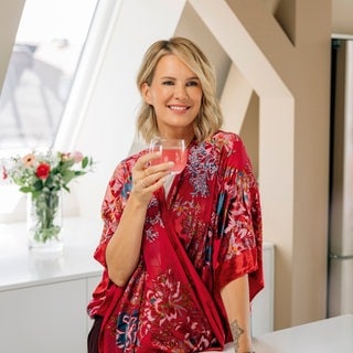 Monica Meier Ivancan steht mit einem Glas an der Hand in einem bunten Wickelkleid in der Küche an der Theke