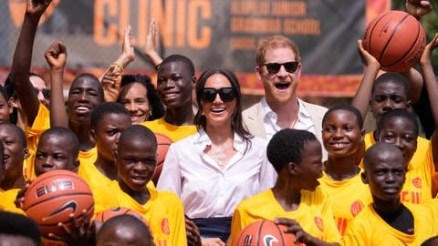 Prinz Harry und Herzogin Meghan stehen in einer Traube Jugendlicher und lachen in die Kamera.