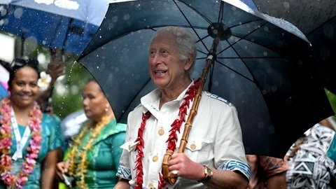 König Charles ist zu Besuch auf Samoa und steht bei Regenwetter unter einem Schirm. Er hat eine Chili-Kette an und lächelt mit den Einwohnern.