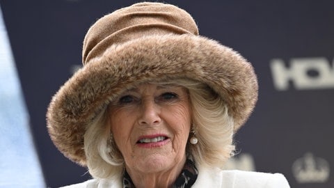 Eine ältere Frau mit blondem Haar und einem großen, hellbraunen Hut mit Kunstfellrand lächelt in die Kamera. Queen Camilla trägt einen hellen Mantel mit Hufeisenbrosche.