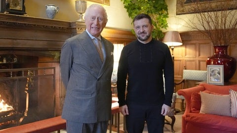 König Charles III. trifft den ukrainischen Präsidenten Wolodymyr Selenskyj auf dem britischen Landsitz Sandringham. Die beiden stehen vor einem Kamin aus Holz und lächeln in die Kamera.