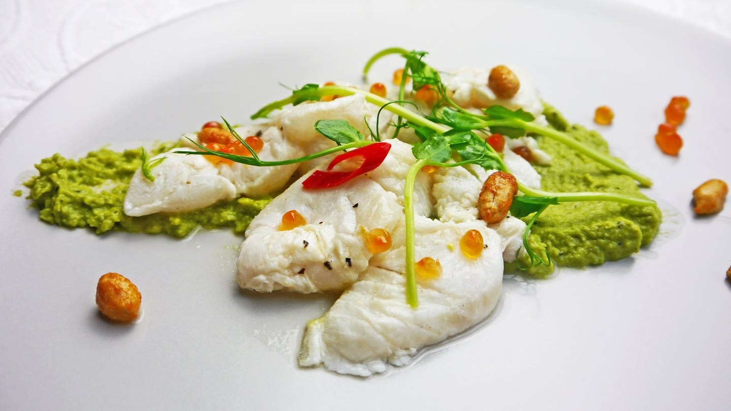 weißer Teller mit grünem Erbsenpüree, darauf helle Fischstücke mit Chili, Nüssen und hellrosa Lachskaviar dekoriert