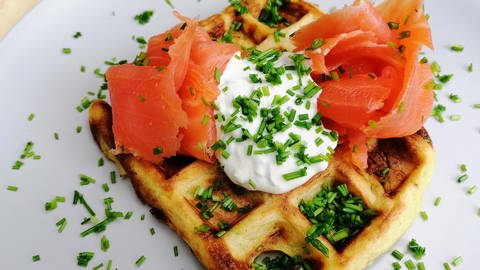 weißer Teller mit quadratischer Waffel mit heller Creme in der Mitte und zwei Lachsrollen rechts und links davon, mit Schnittlauch bestreut