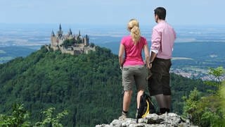 Zwei Wanderer blicken vom Zeller Horn der Schwäbischen Alb auf die Burg Hohenzollern bei Hechingen. 