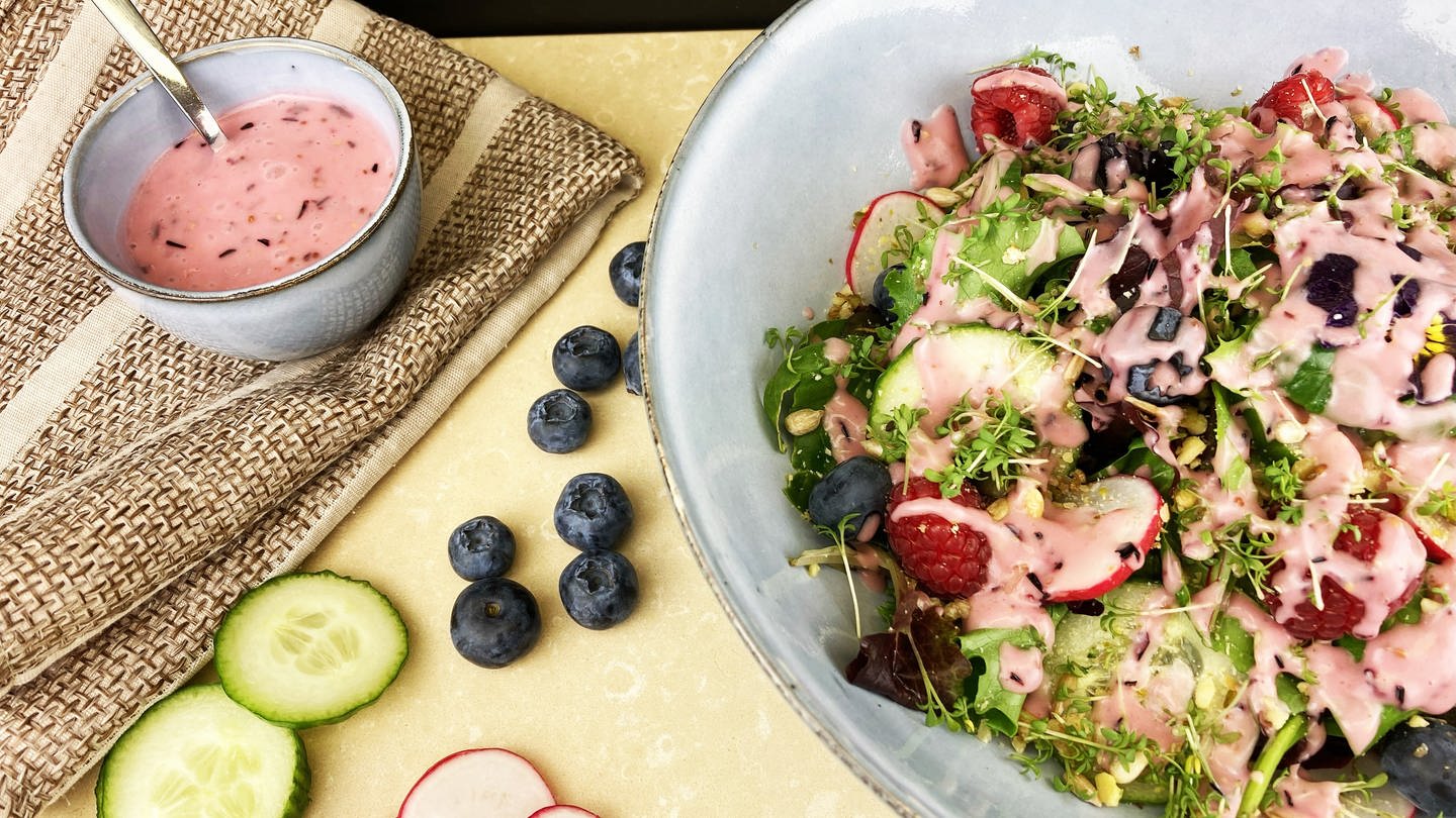 Schüssel mit Salat mit rosa Soße, daneben Blaubeeren, Gurkenscheiben und ein Töpfchen mit rosa Dressing